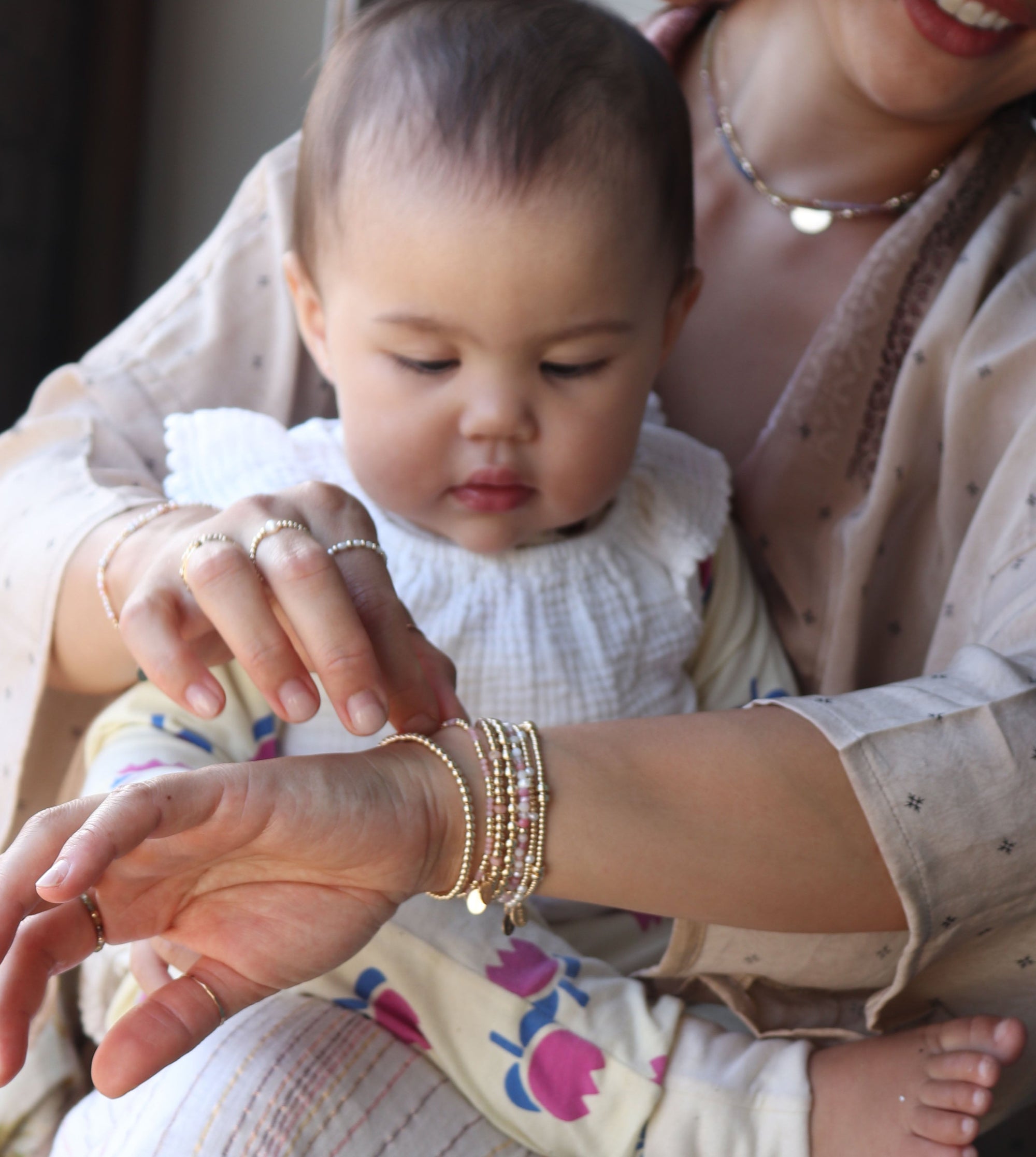 Mother holding her baby, wearing multiple pieces of jewellery with pink gemstones and pearls, rings, bracelets and necklaces.