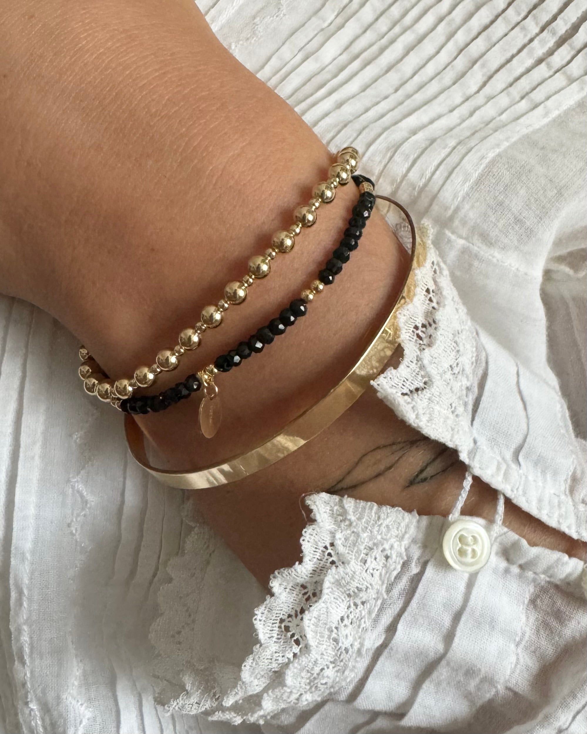 Close-up of a wrist wearing a gold beaded bracelet layered with a black spinel gemstones gold beaded bracelets and a gold bangle with a lace white top