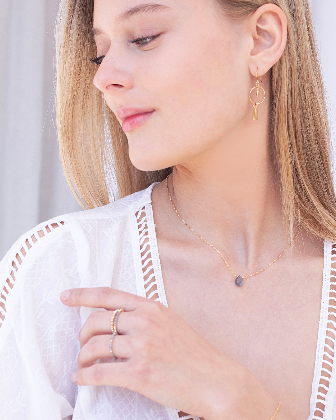 Woman wearing a gold necklace with a labradorite tear drop gemstone, gold earrings and rings, with a blurred background