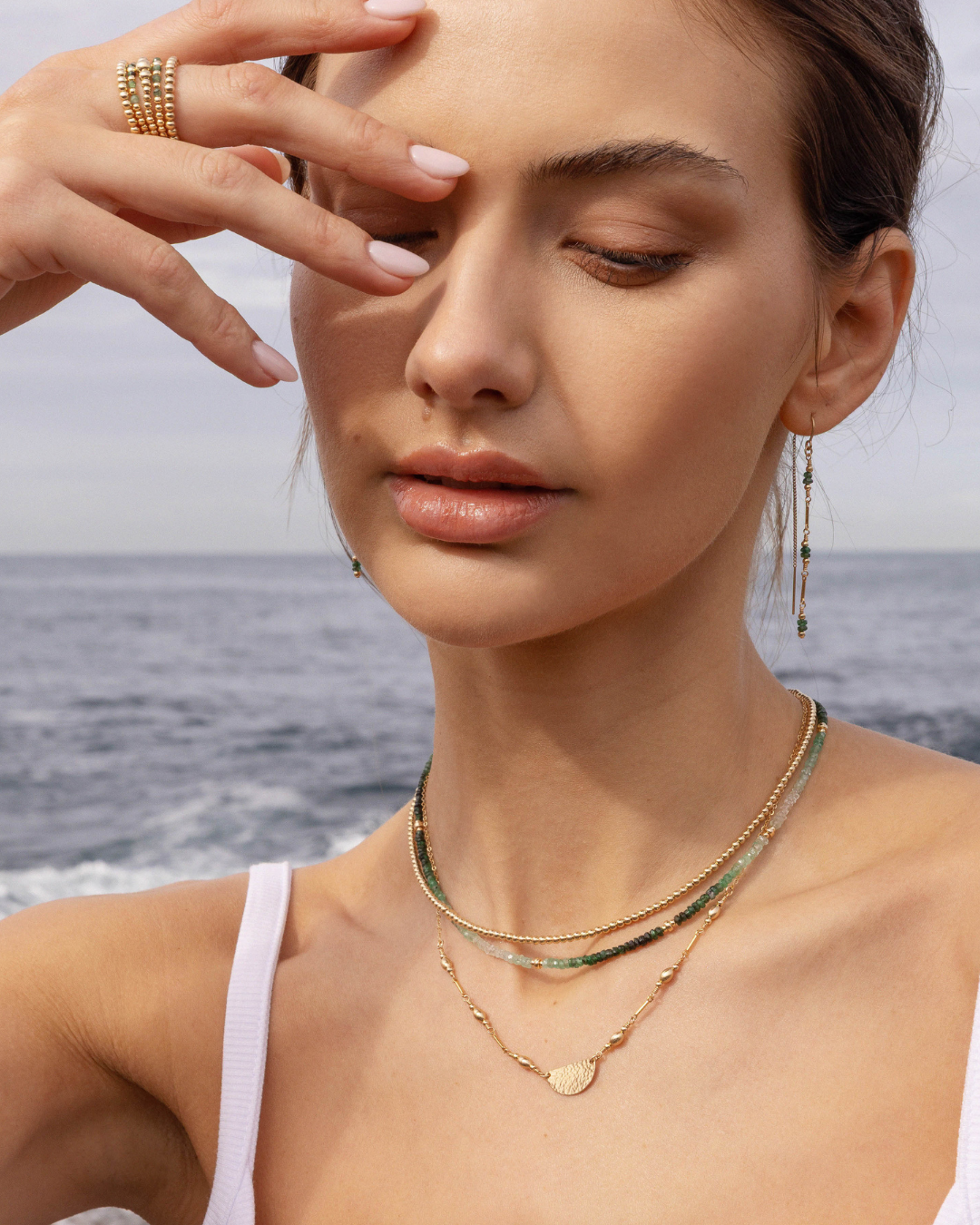 Woman wearing layered gold necklaces and green emerald bead earrings with a blurred ocean background