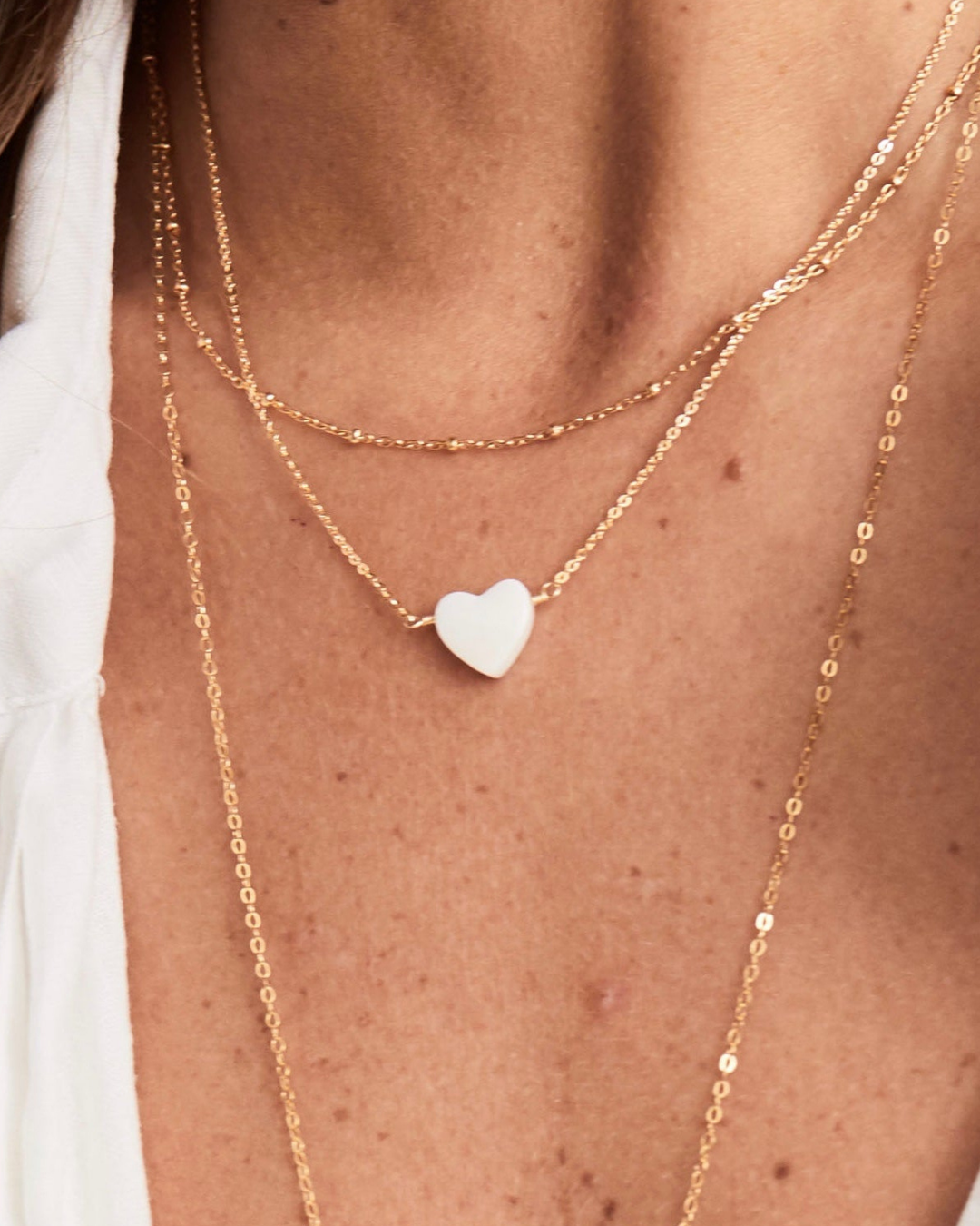 Close up of neck with gold necklace with a heart shaped mother of pearl pendant layered with other gold chains.