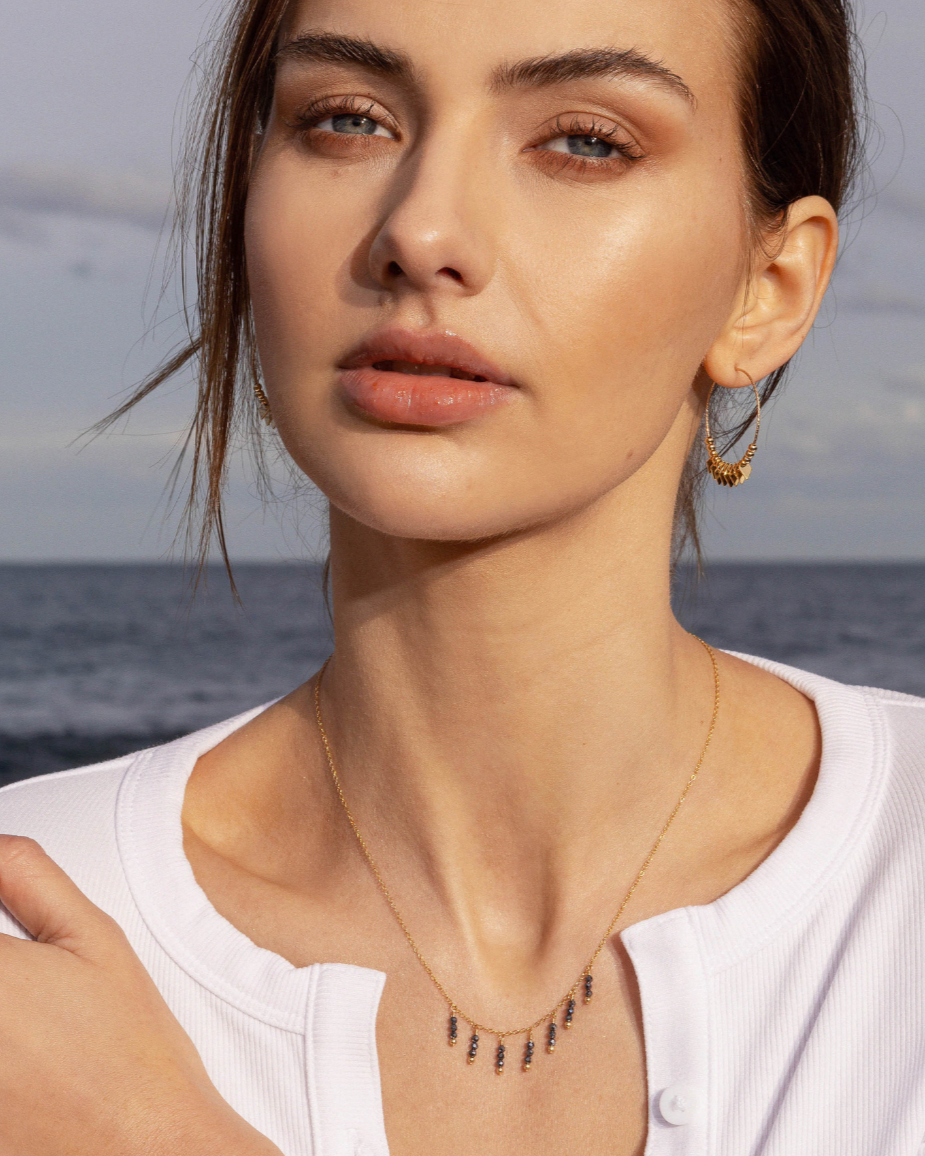 Woman wearing gold hoop earrings and a black gemstone necklace with a blurred ocean background