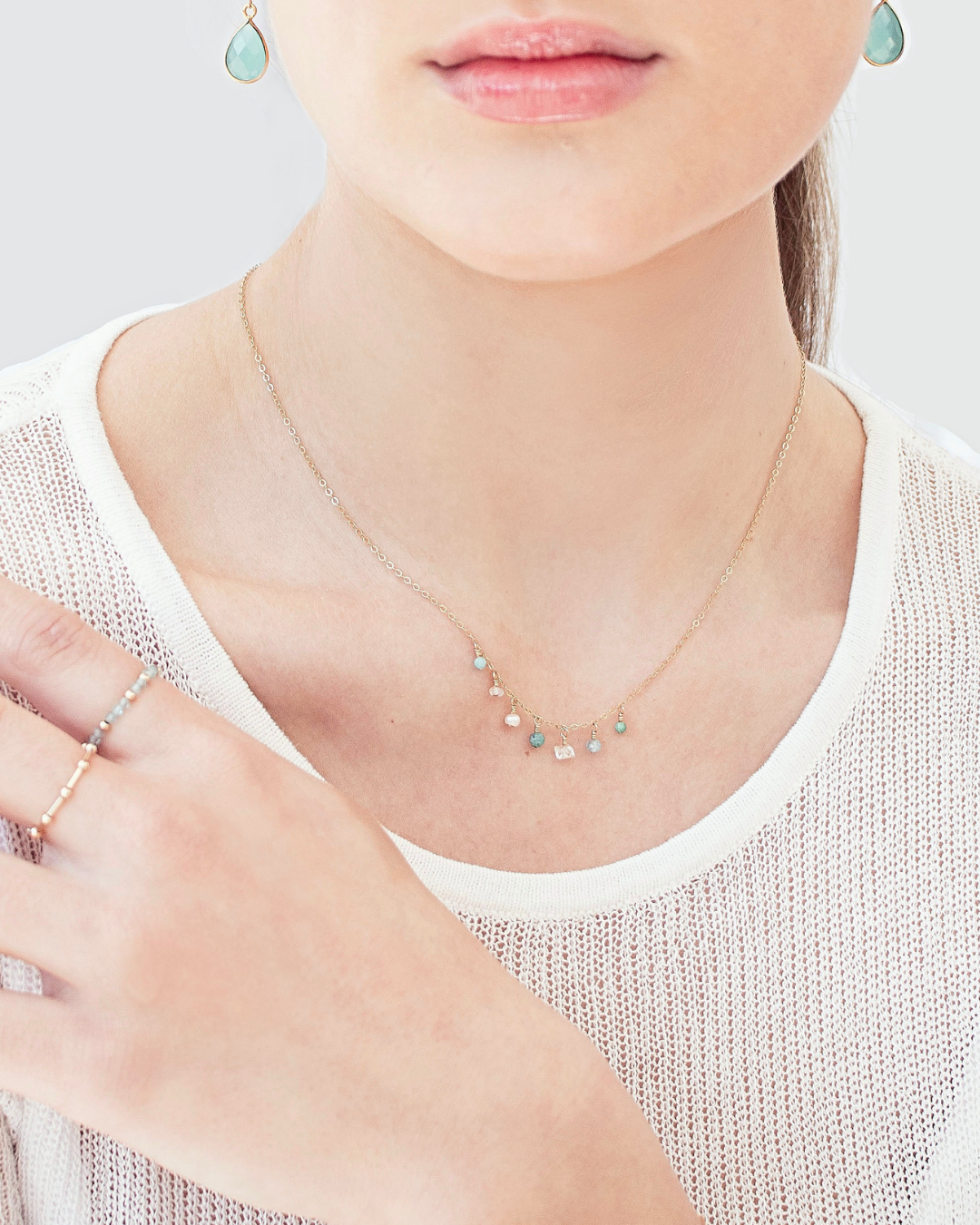Close-up of a person wearing a delicate necklace with small gemstones and pearl, chalcedony earrings, and gold rings.