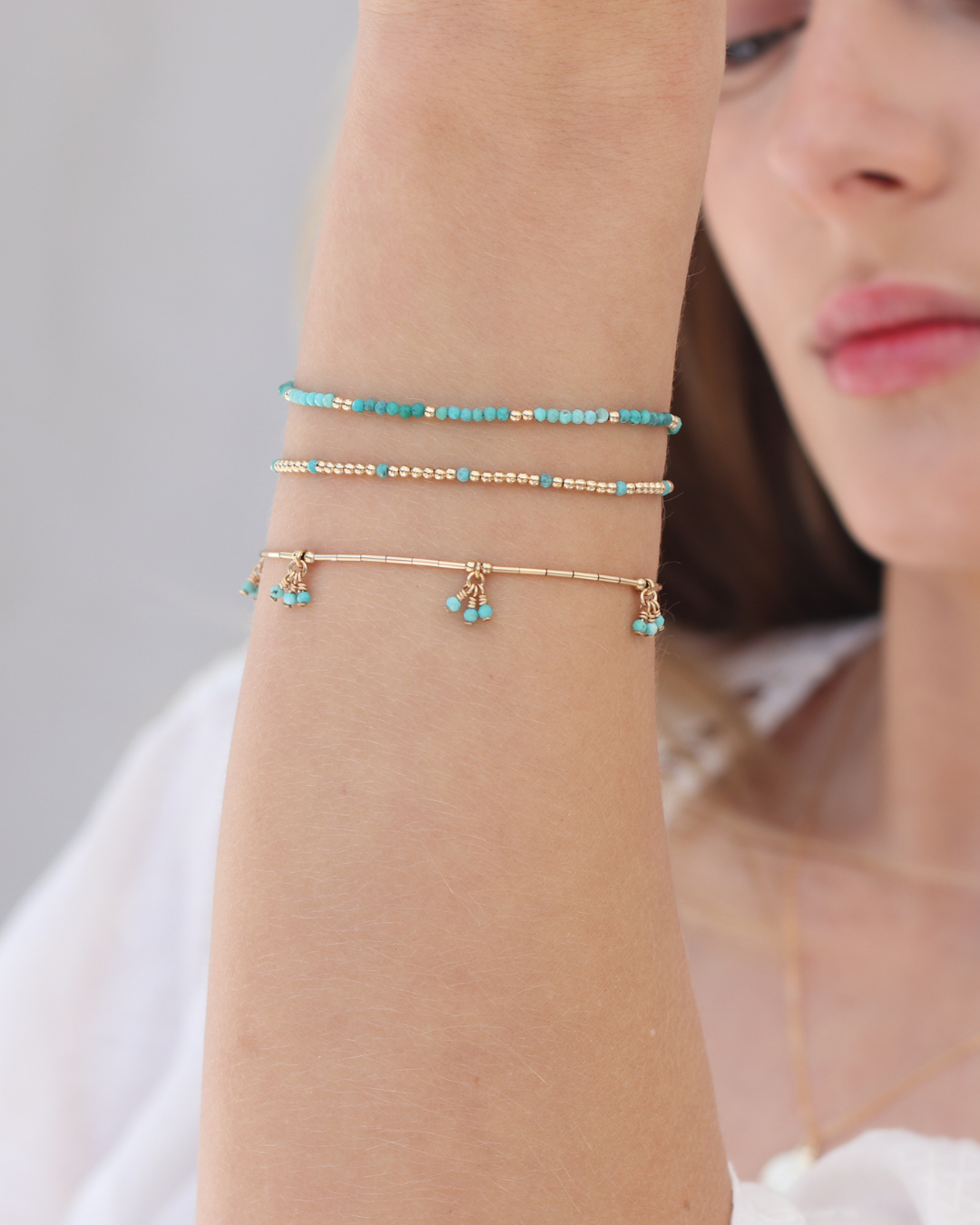 Close-up of a person wearing gold and turquoise gemstone bracelets on their wrist on a neutral background