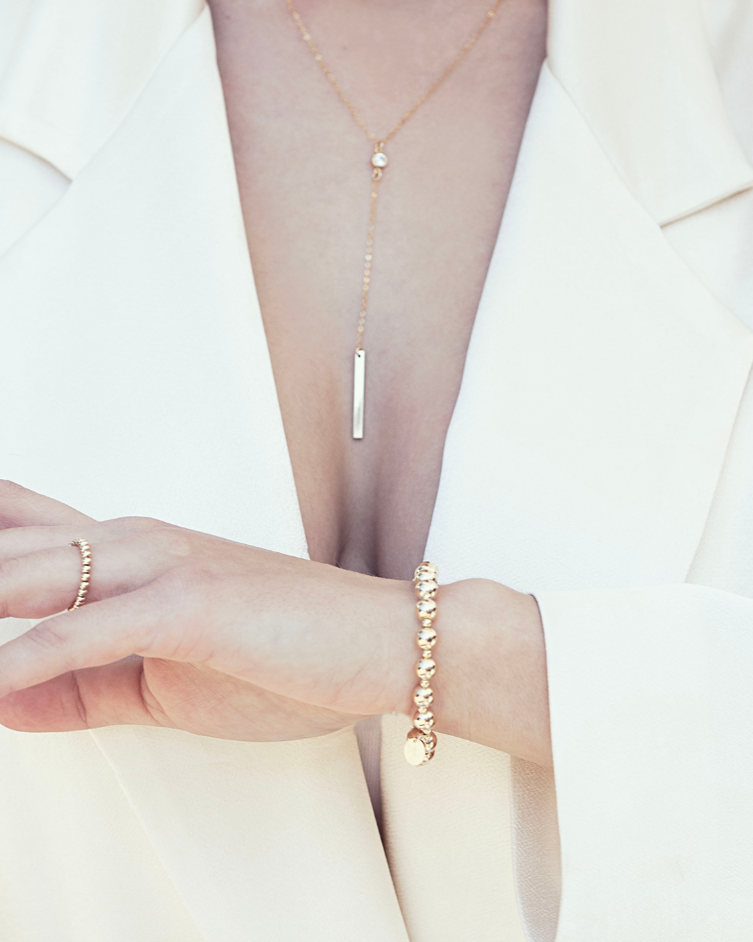 Model wearing a gold bead bracelet, ring and fine gold lariat cubic zirconia gemstone necklace, bracelet, wearing a cream satin outfit.