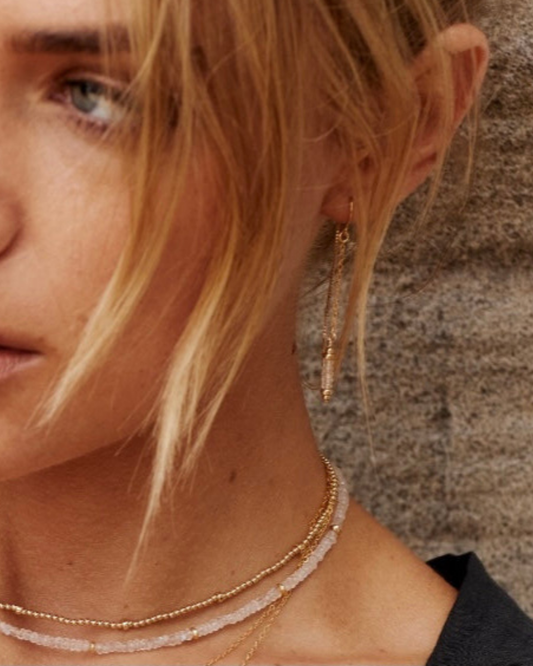 Close-up of a person wearing gold and moonstone gemstone earrings and necklaces against a neutral background