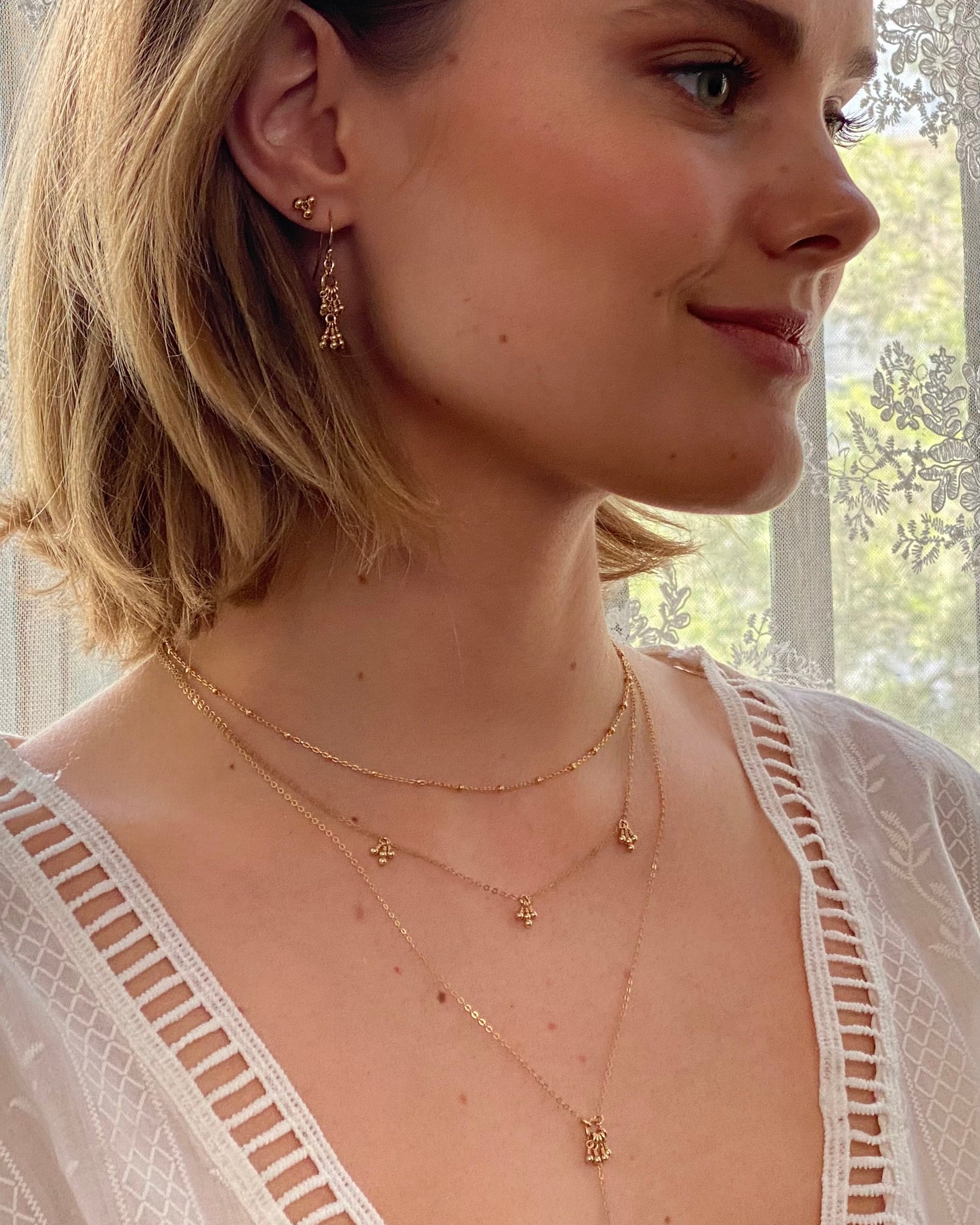 Woman wearing layered gold delicate necklaces and earrings with a blurred outdoor background