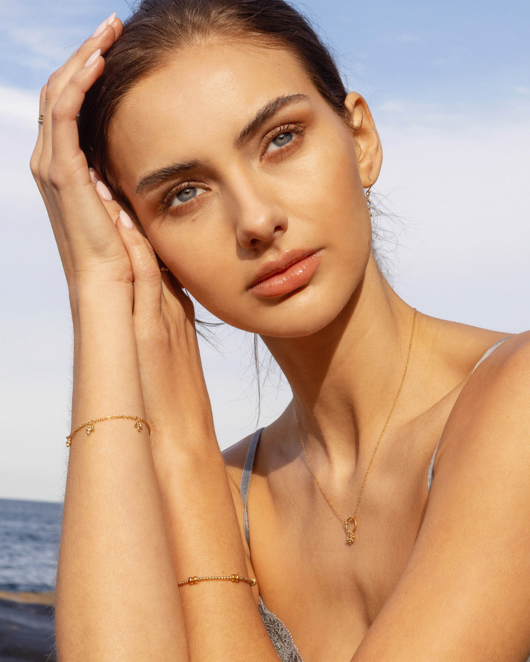 Woman wearing delicate gold jewellery including a necklace, bracelets and earrings against a clear blue sky
