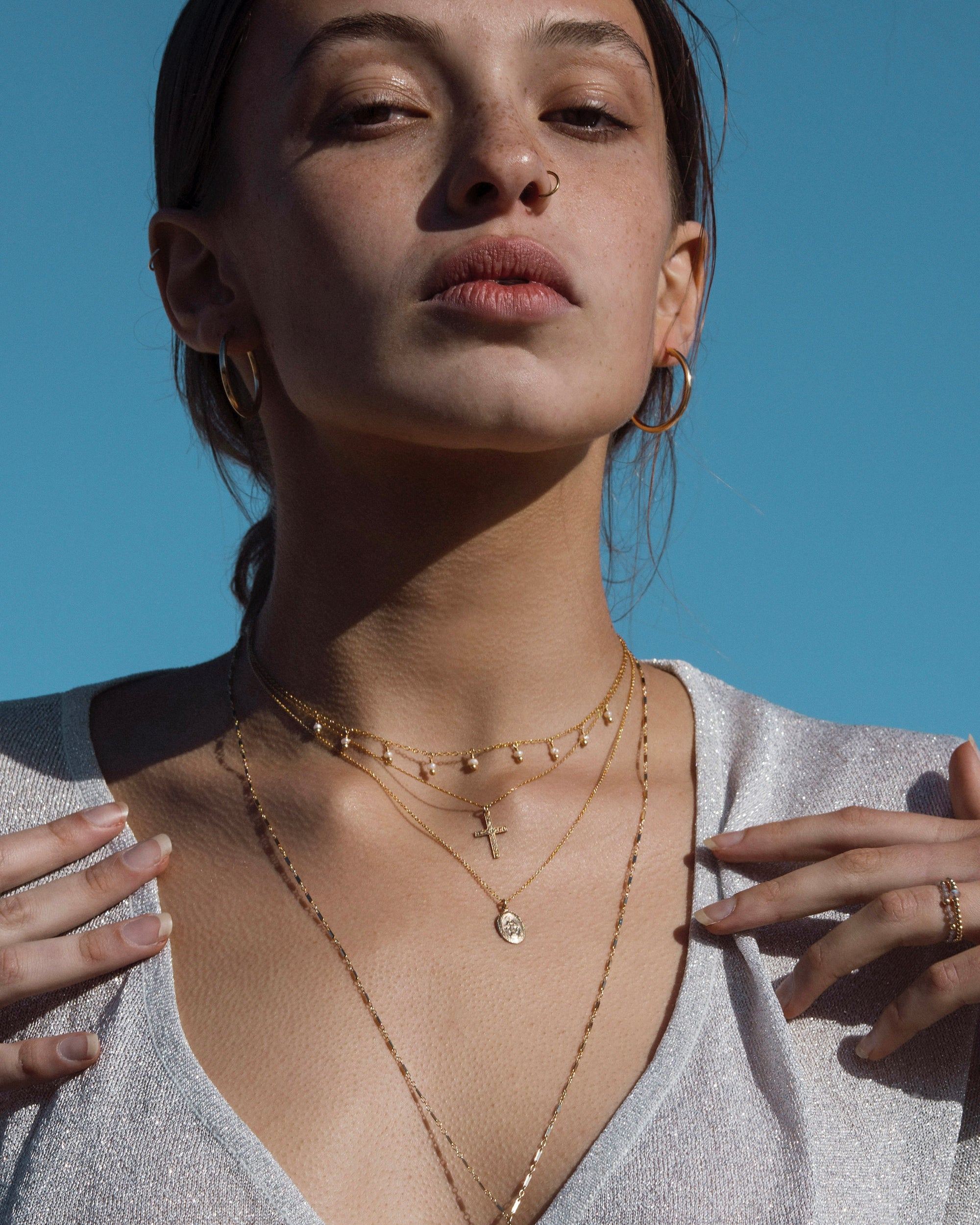 Woman wearing gold hoop earrings, rings and a gold cross pendant necklace layered with multiple gold necklaces against a clear blue sky