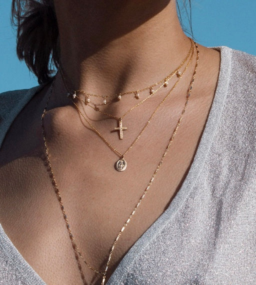 Close-up of woman's neck wearing a mary coin necklace layered with a cross pendant necklace and other gold necklaces with a blue sky in the background.