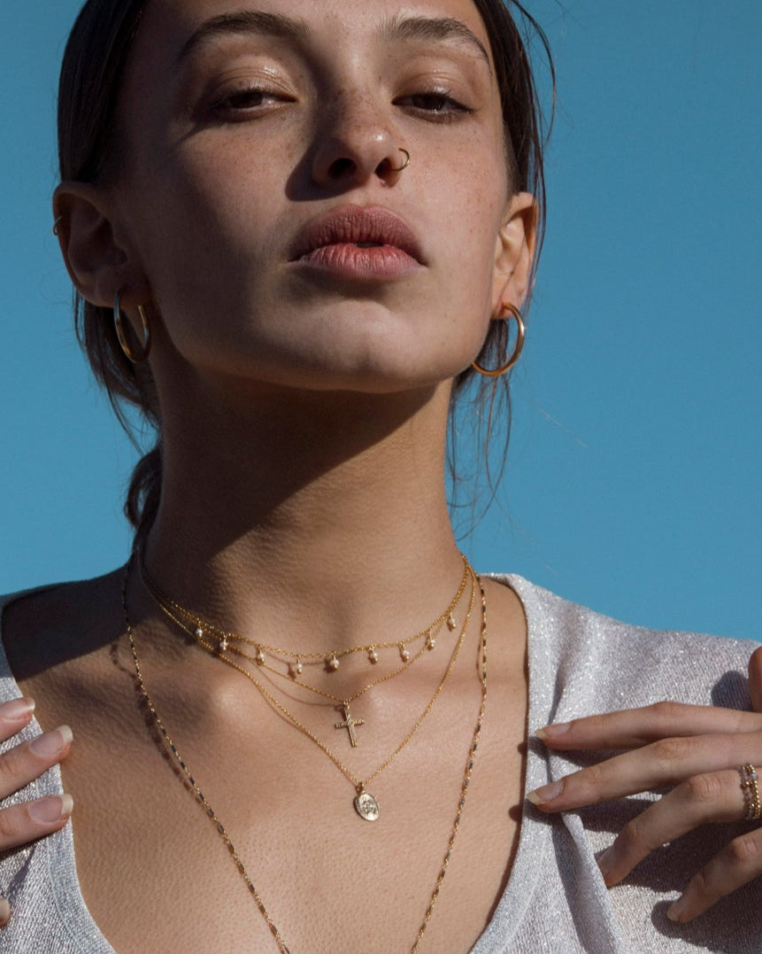 Woman wearing gold hoop earrings and necklaces featuring a Mary and cross pendant against a clear blue sky.