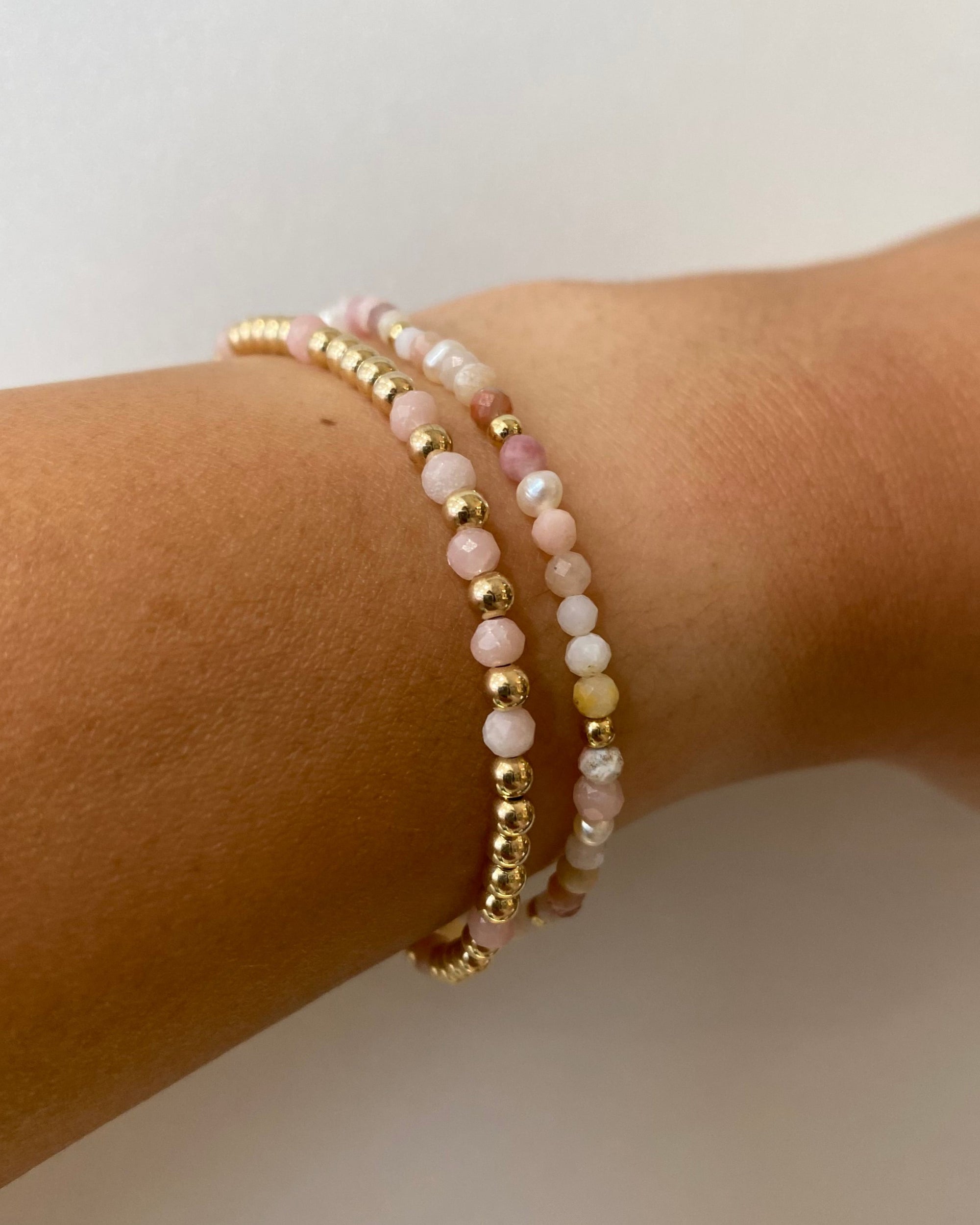 Two gold bead bracelets with pink gemstones and pearl on a wrist against a neutral background