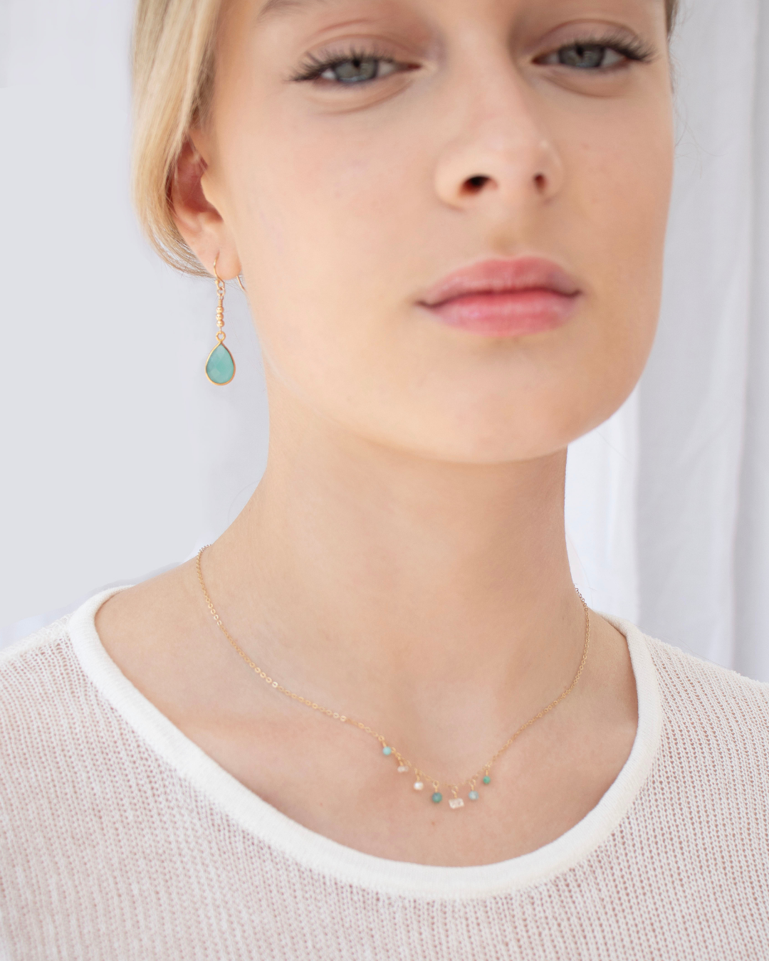 Close-up of a person wearing a delicate necklace with small aqua gemstones and a pair of Chalcedony earrings.