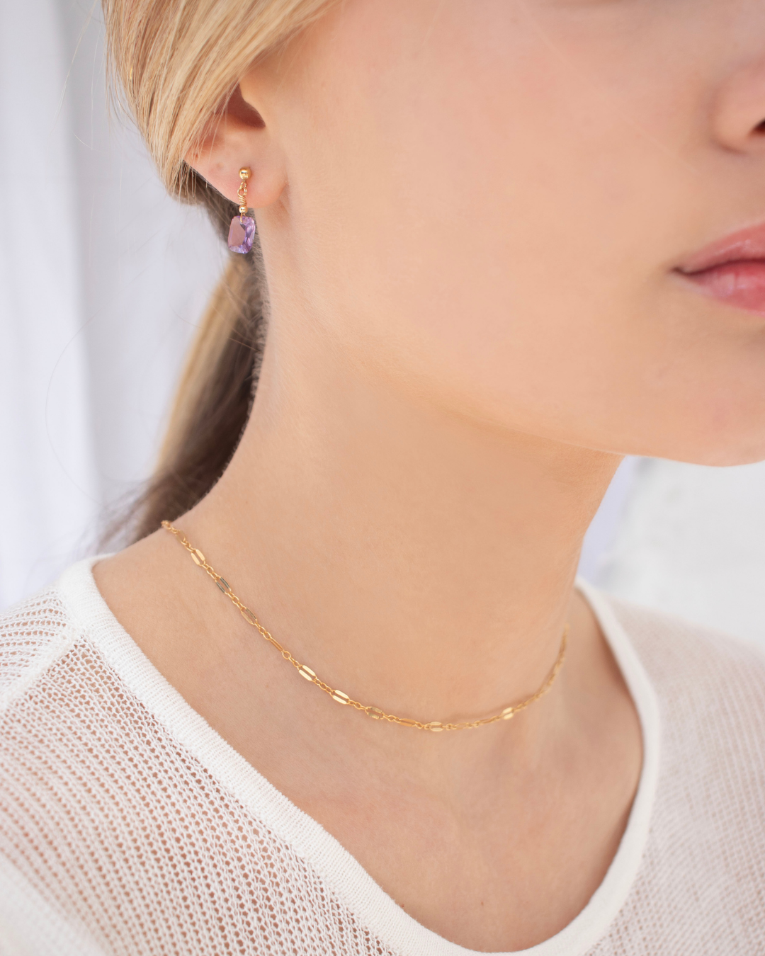 Close-up of a person wearing purple amethyst gold earrings and a gold choker necklace with a blurred background
