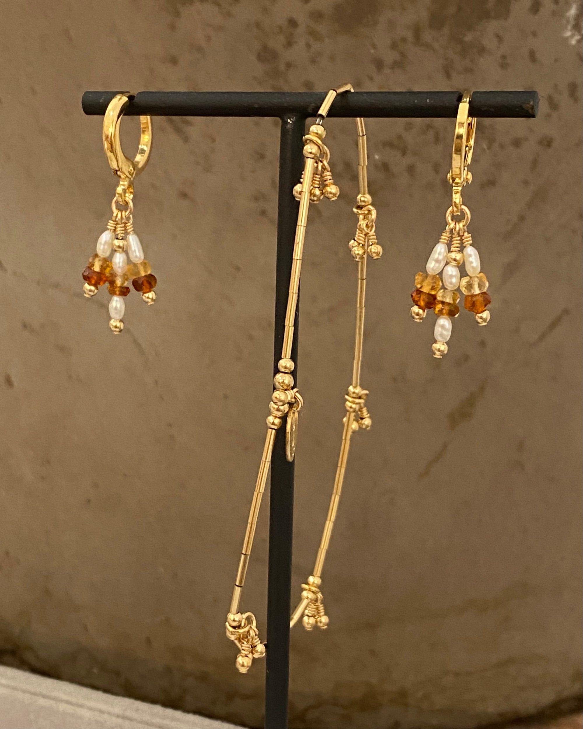 Gold hoop earrings with citrine gemstones and pearls dangling on a black stand against a brown background