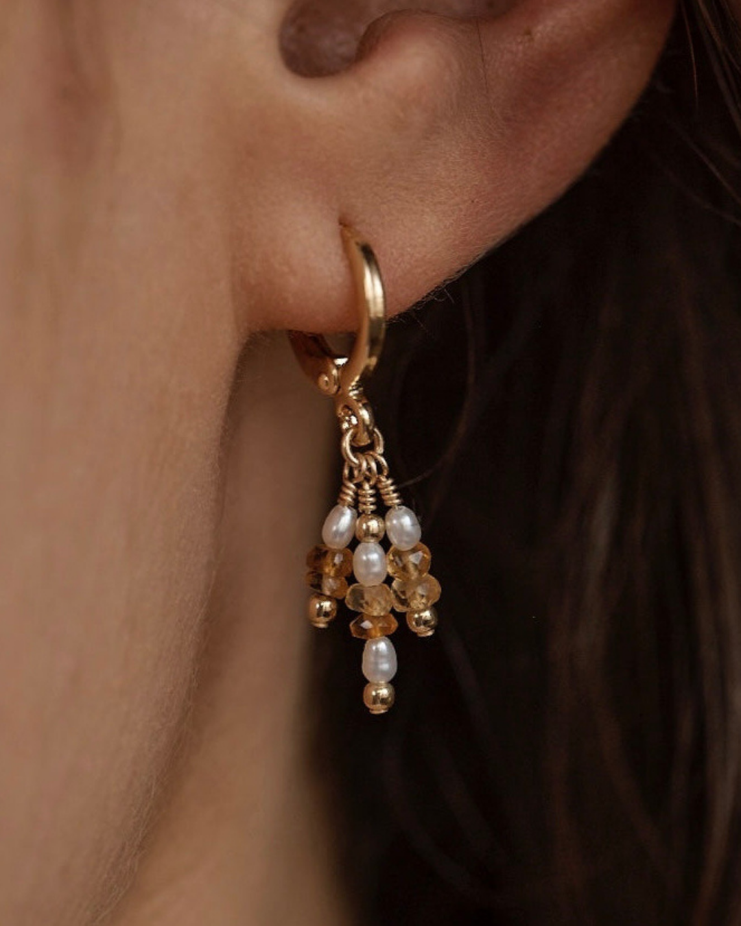 Close up of gold hoop earring with pearl and citrine gemstone drops worn by a person.