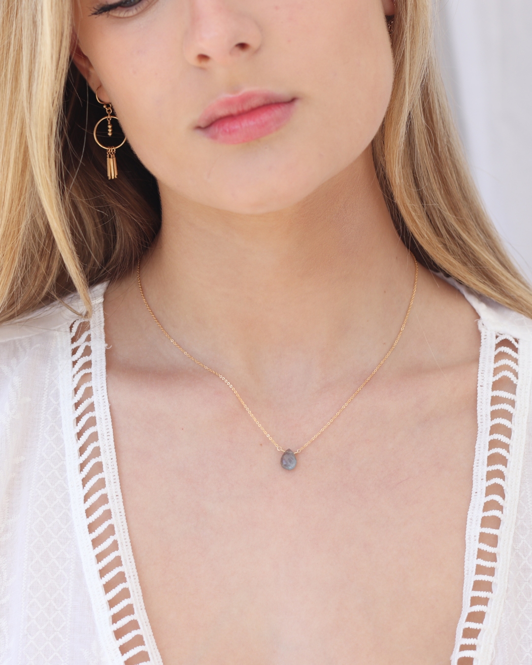 Close-up of a person wearing a delicate necklace with a teardrop-shaped labradorite pendant on a gold chain against a white background
