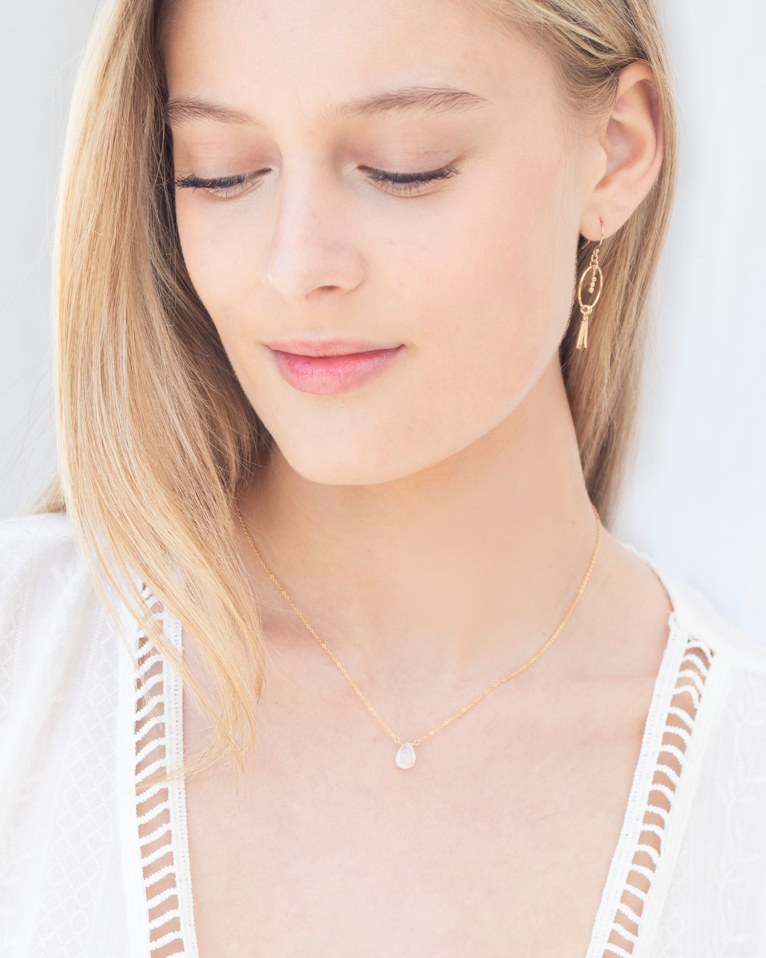 Woman wearing a gold delicate moonstone necklace and gold earrings on a plain background