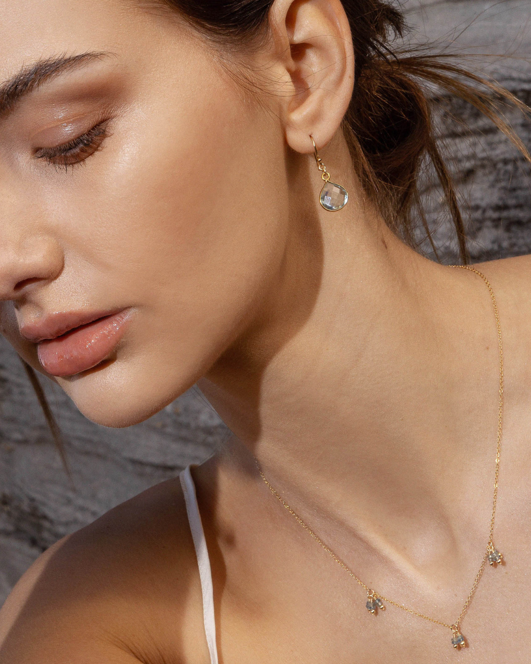 Close-up of a woman wearing gold earrings and necklace with aquamarine gemstones against a stone wall background