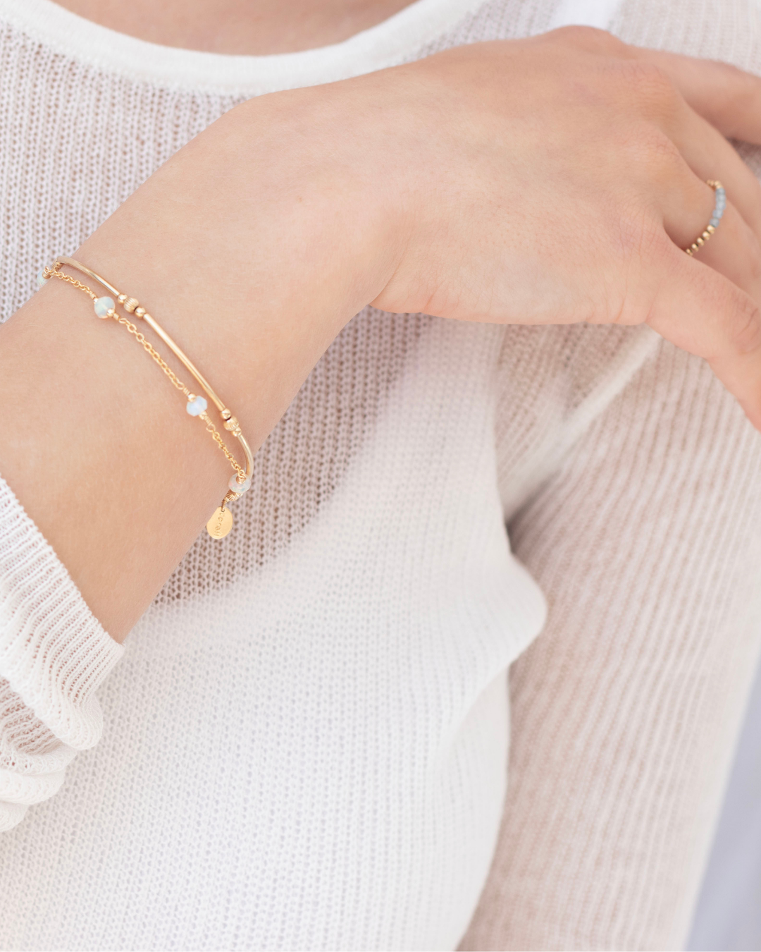 Close-up of a models hand wearing a ring and multiple gold bracelets on a light background