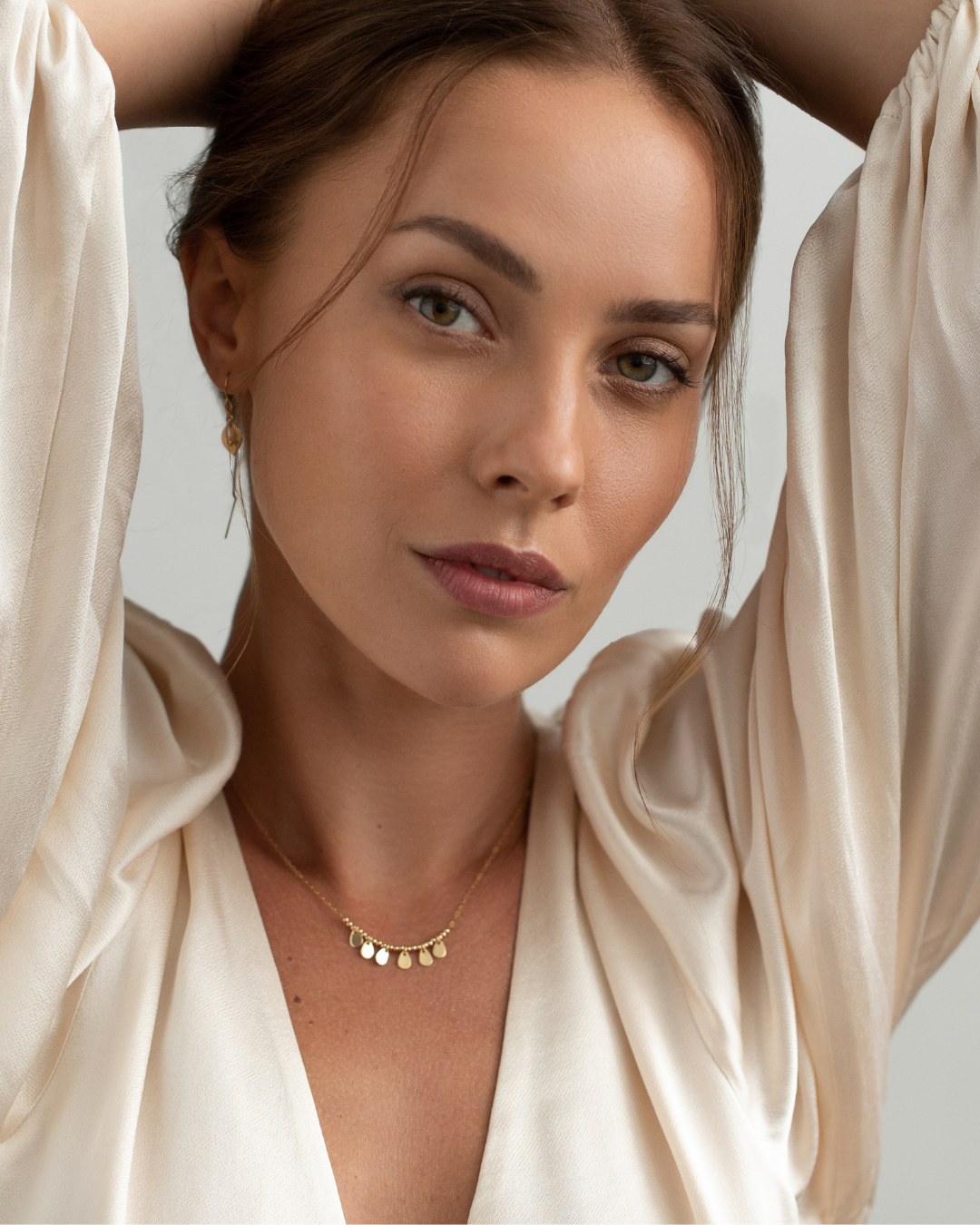 Woman wearing a gold necklace with teardrop gold pendants and a white silk blouse on a plain background