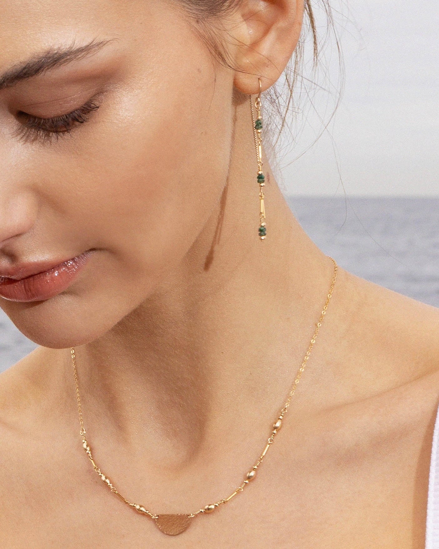 Close-up of a woman wearing a gold necklace and emerald bead earrings with a blurred ocean background