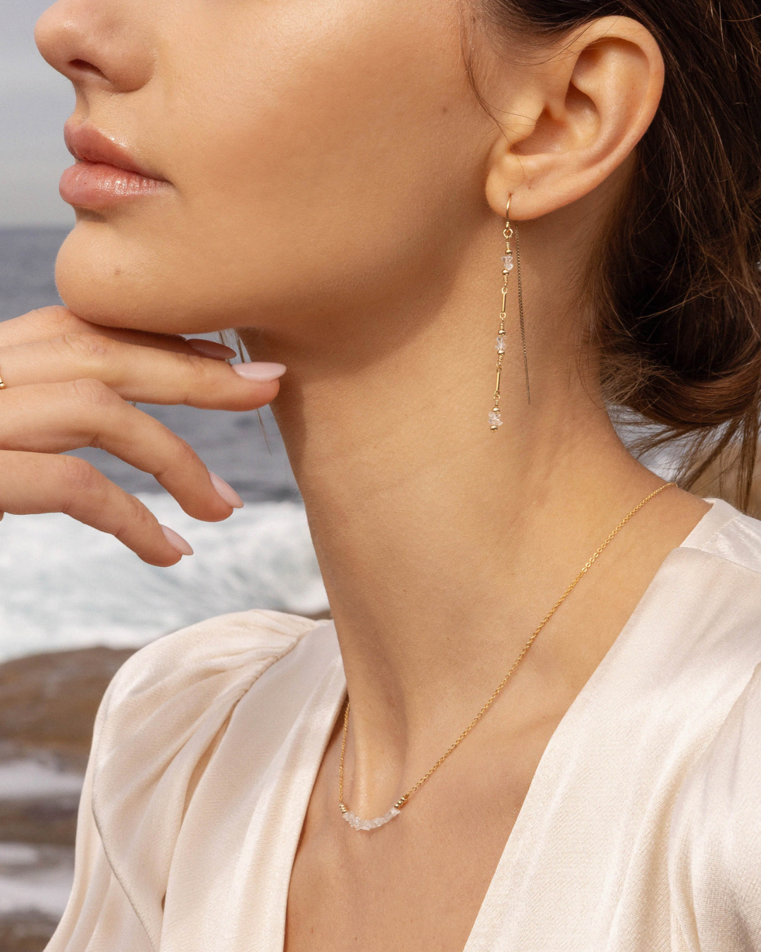 Close-up of a woman wearing gold and herkimer diamond earrings and a necklace with a blurred ocean background