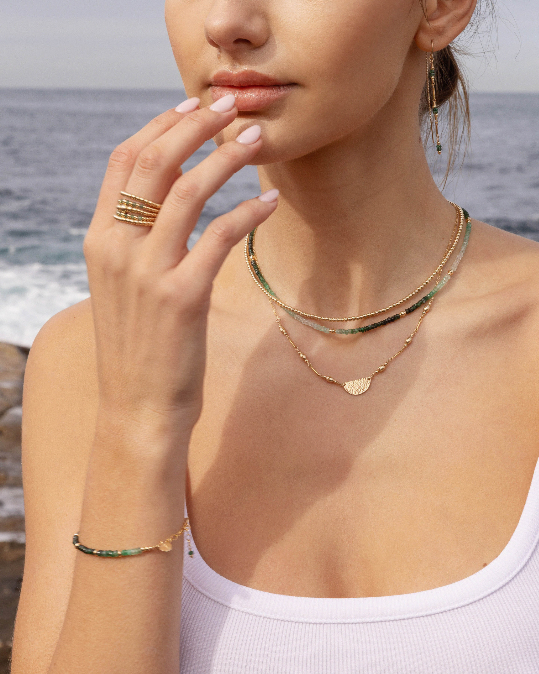 Woman wearing layered gold and beaded jewellery with ocean background.