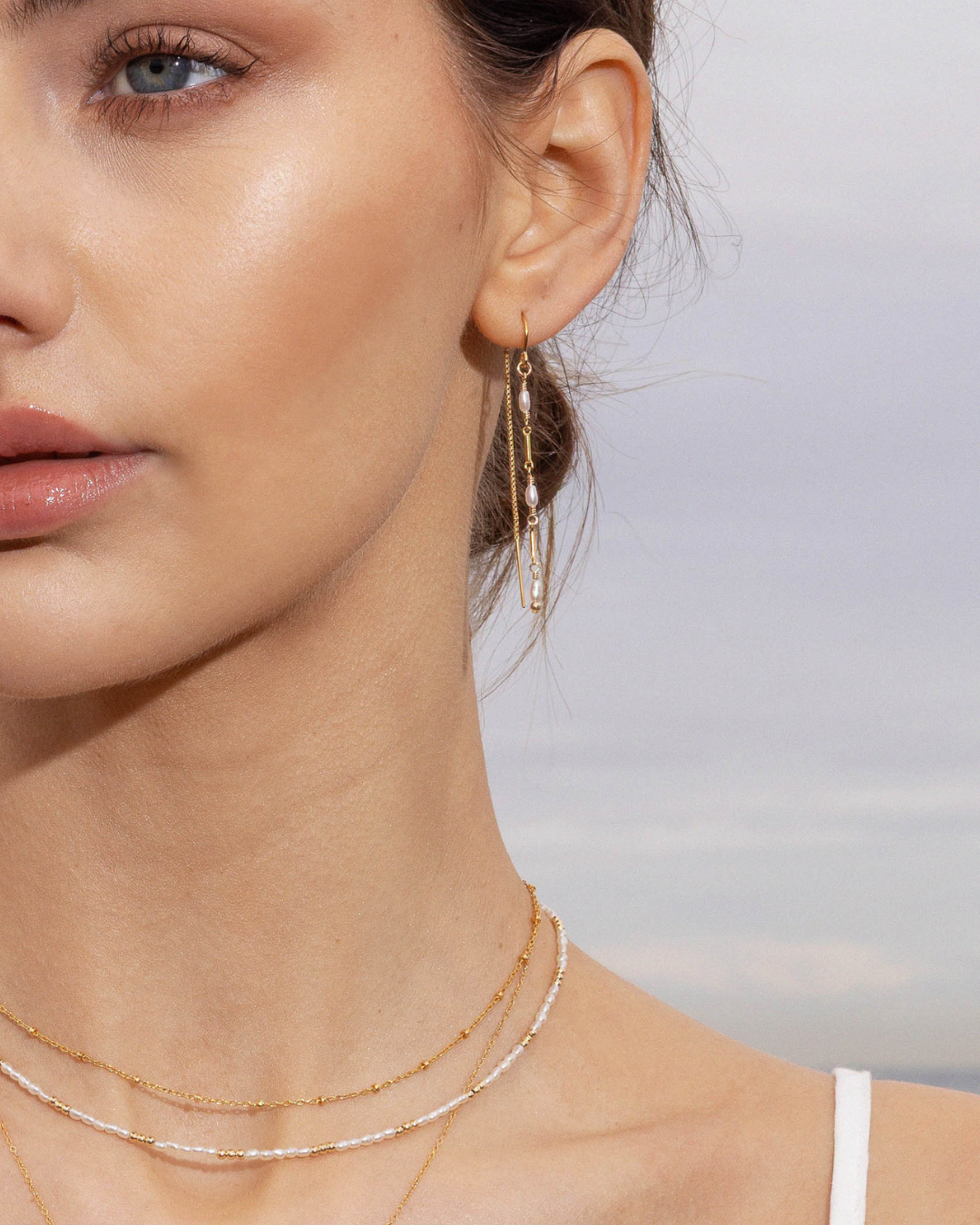 Close-up of a woman wearing gold pearl beaded earrings and necklaces against a blurred natural background