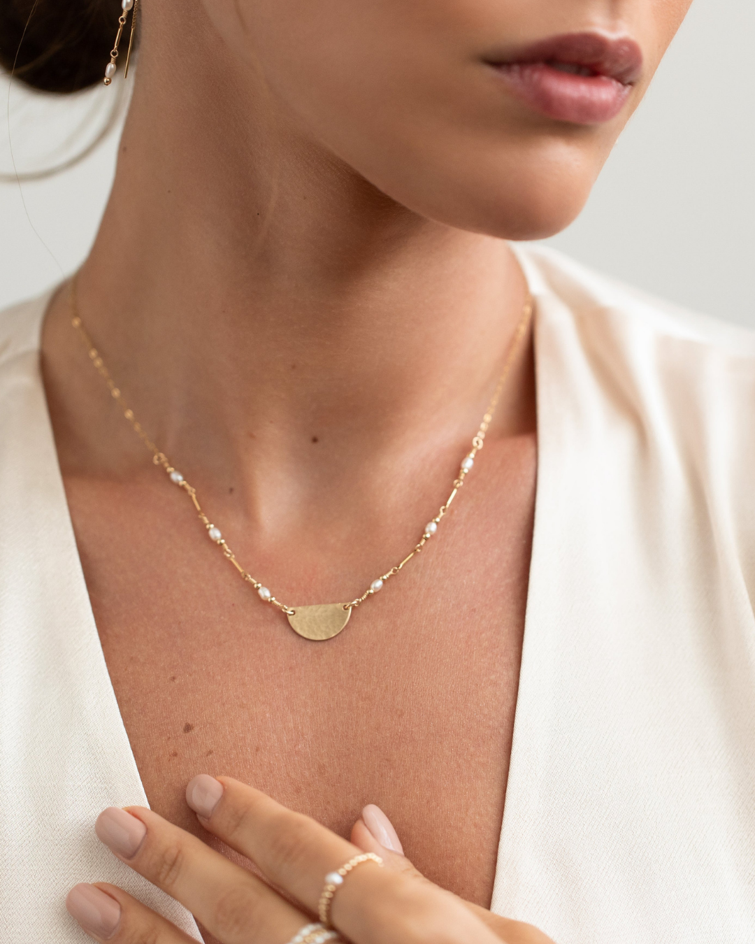 Close-up of a person wearing a gold necklace with pearls and a half disc pendant, with a blurred background.