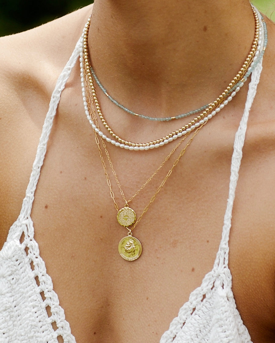 Close-up of a person wearing multiple gold pendant necklaces with a pearl and apatite necklace in a white crochet bikini top.