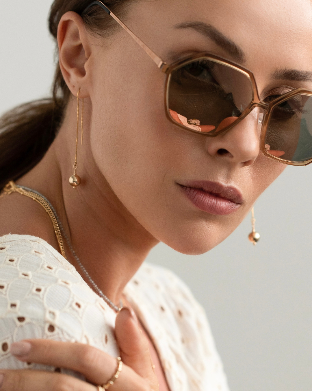 Woman wearing gold sunglasses and jewelry against a neutral background