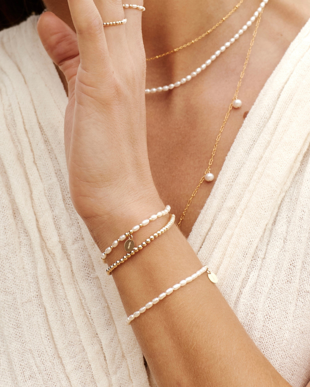 Close-up of a person wearing multiple pearl and gold necklaces and bracelets on a neutral background