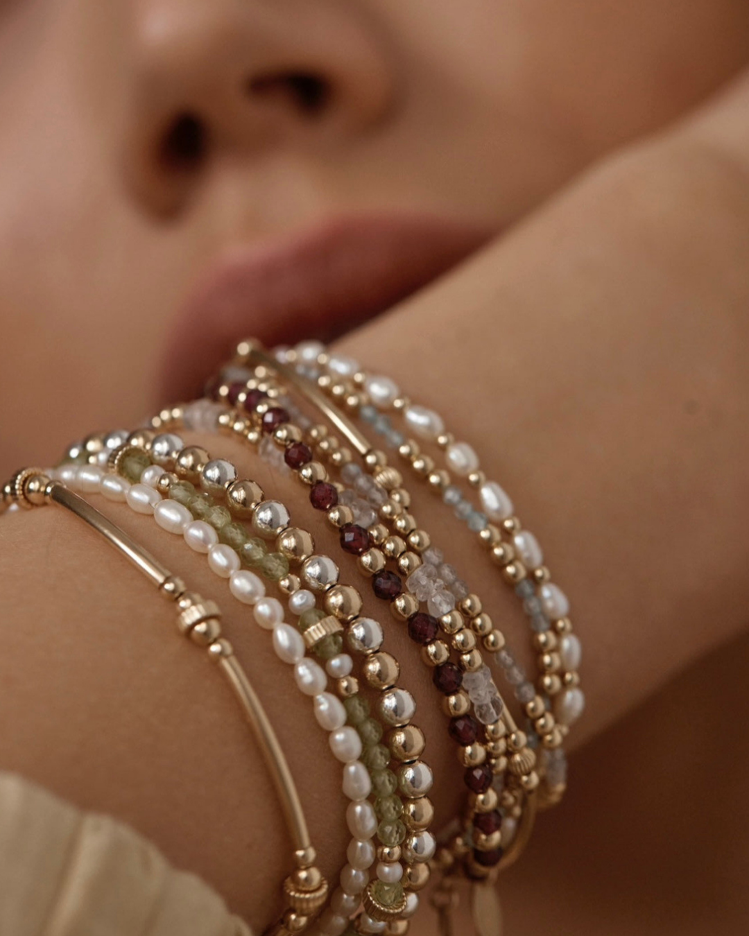 Close-up of a person's wrist wearing multiple beaded gold and pearl bracelets on a blurred background