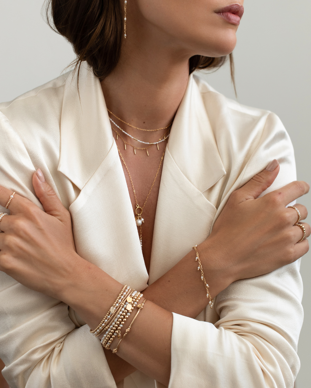 Woman wearing gold and pearl jewellery including necklaces, bracelets, and rings on a neutral background