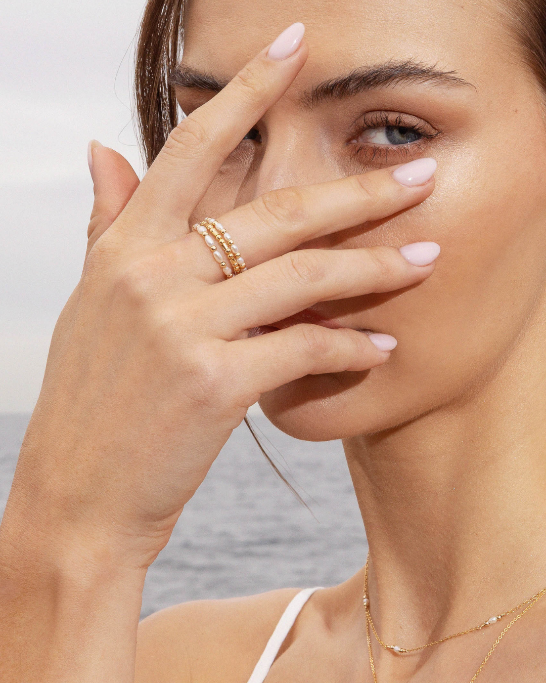 Close-up of a woman's hand with pink nail polish and pearl and gold rings, touching her face against a blurred background.