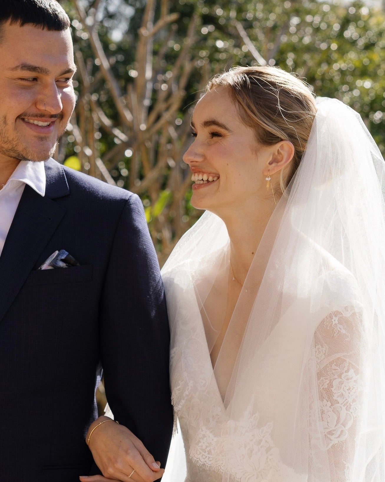 Bride with cubic zirconia jewellery and her groom in wedding attire standing close together outdoors with trees in the background.