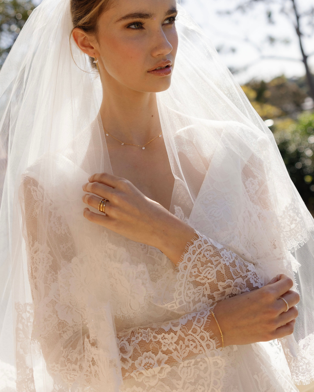 Bride wearing a lace wedding dress and veil outdoors with gold delicate jewellery.