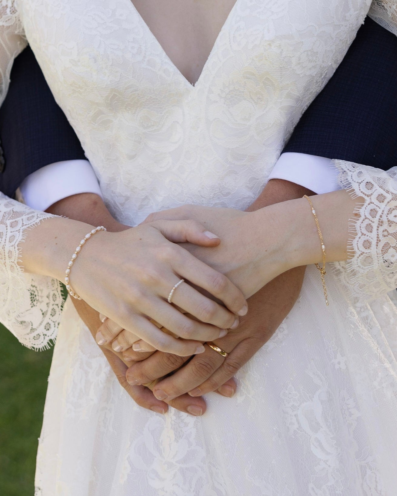 Close-up of bride and grooms hands holding each other with wedding rings, wearing pearl and gold bracelets, against a blurred background.
