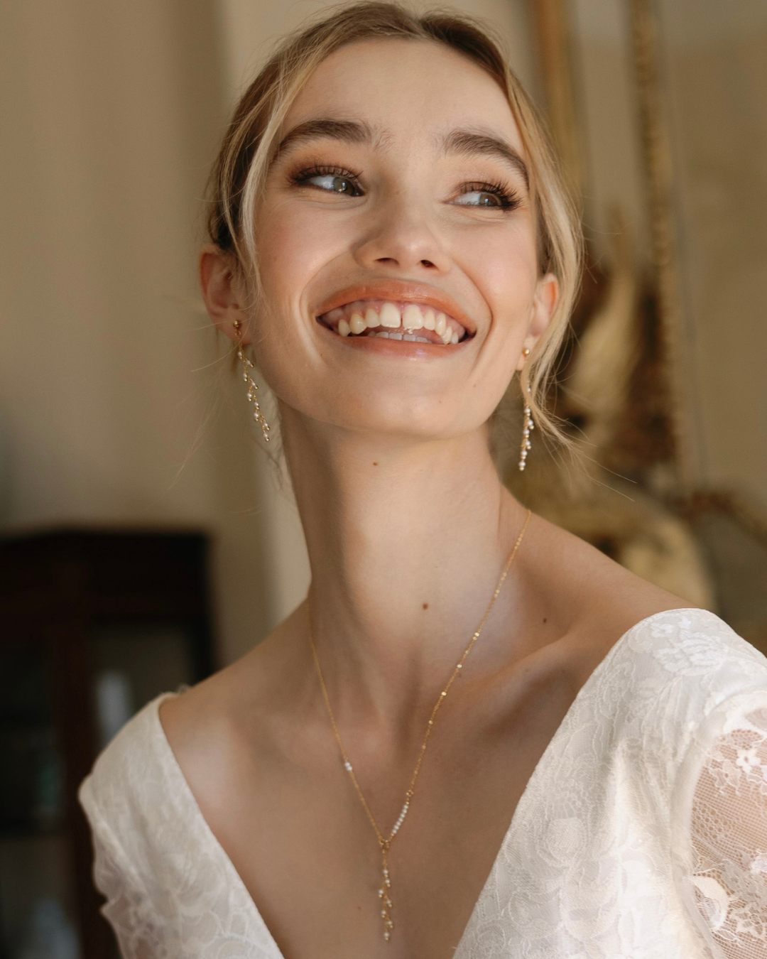 Bride wearing a gold necklace and gold and pearl earrings, smiling in a softly blurred indoor setting