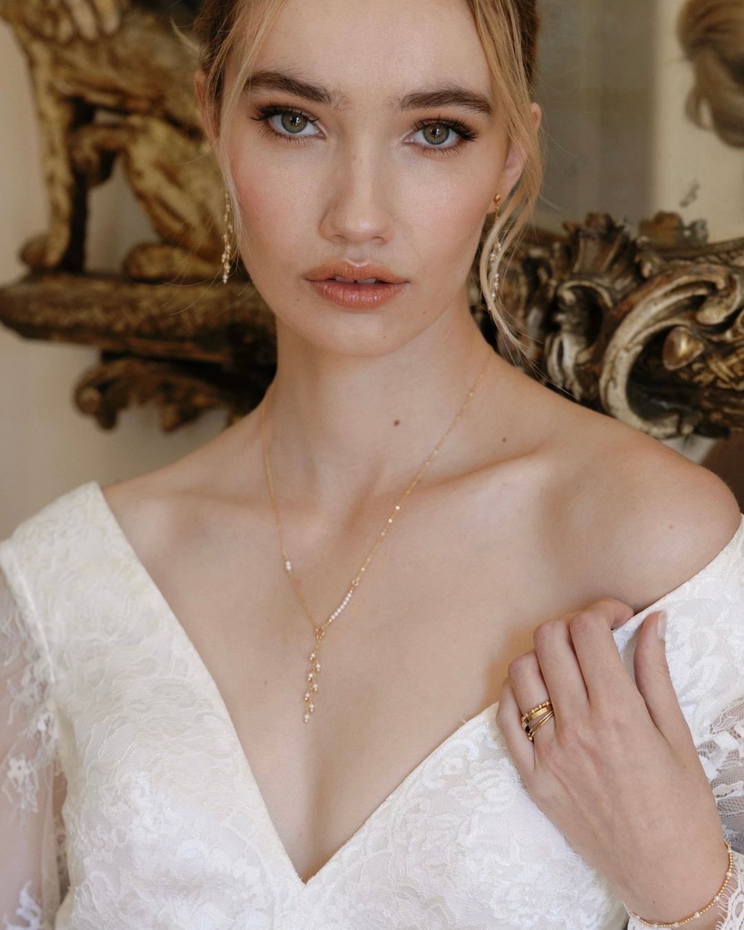 Bride wearing a gold and pearl necklace and rings in front of an ornate mirror.