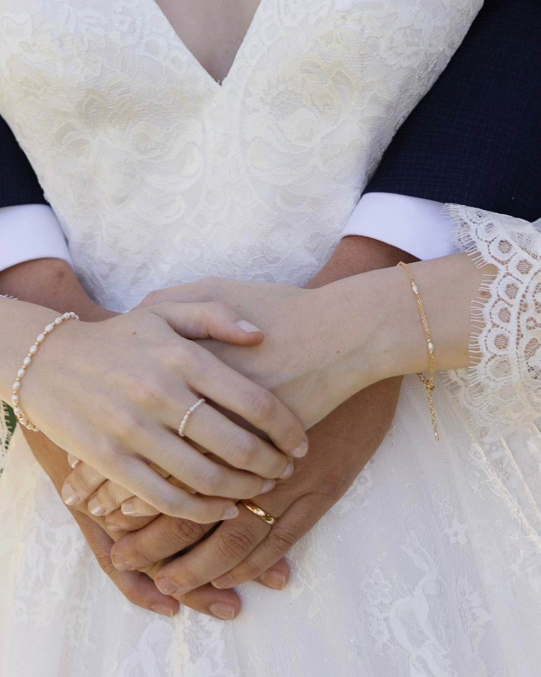 Close-up of hands of bride and groom holding each other with wedding rings and gold and pearl bracelets.