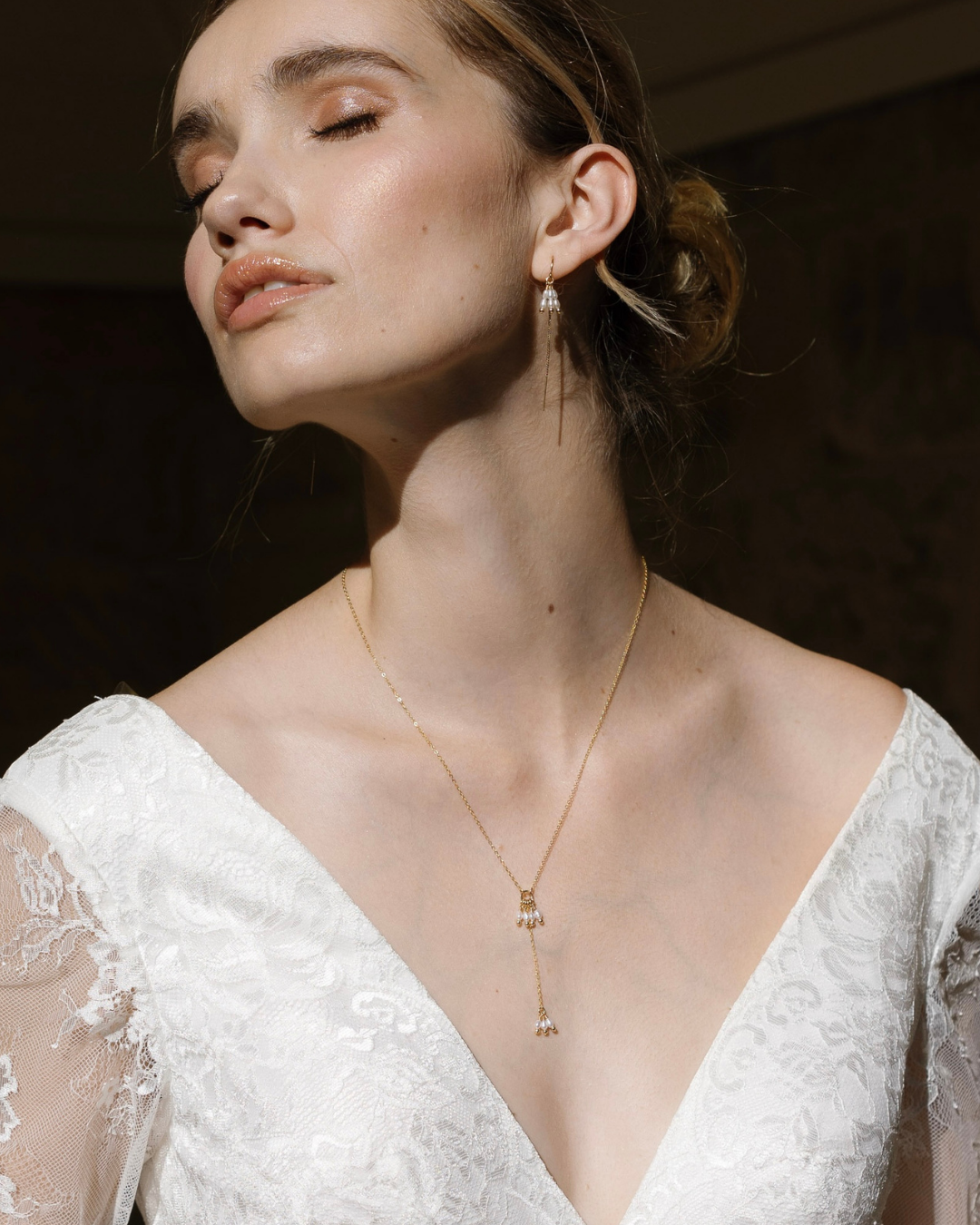 Bride in wedding dress wearing a delicate gold and pearl necklace set against a dark background.