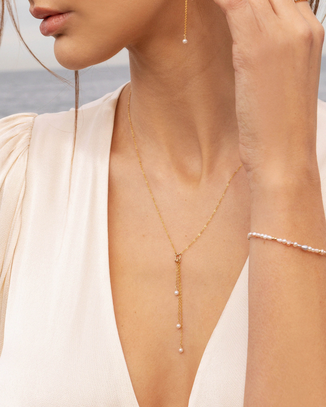 Close-up of a person wearing a delicate gold lariat necklace with pearl drops against a neutral background.