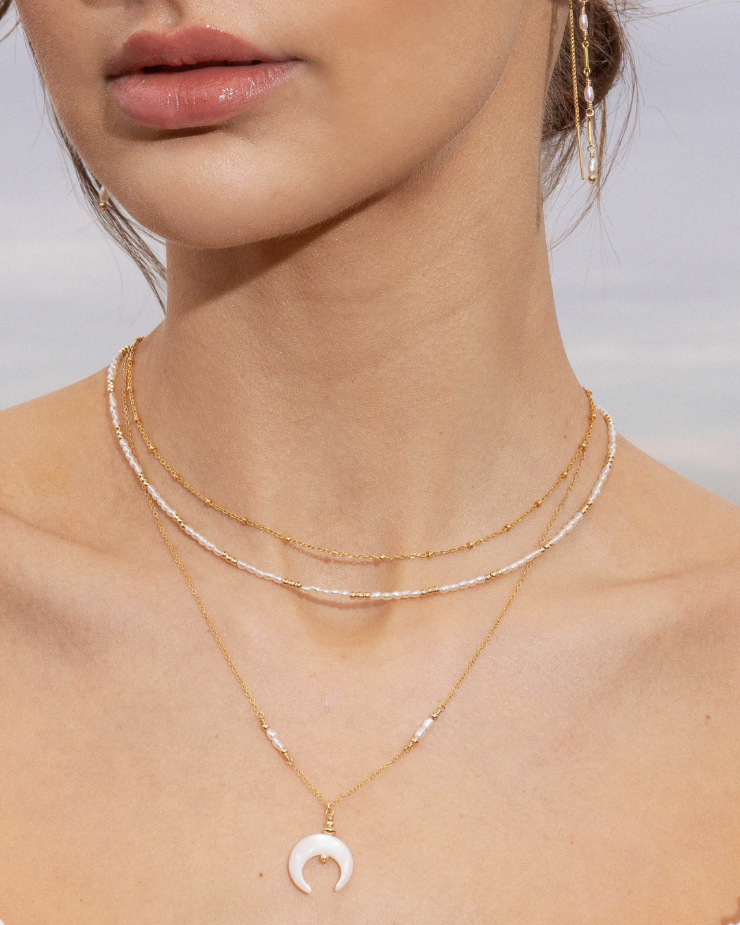 Close-up of a person wearing layered pearl and gold necklaces with pearl earrings on a neutral background