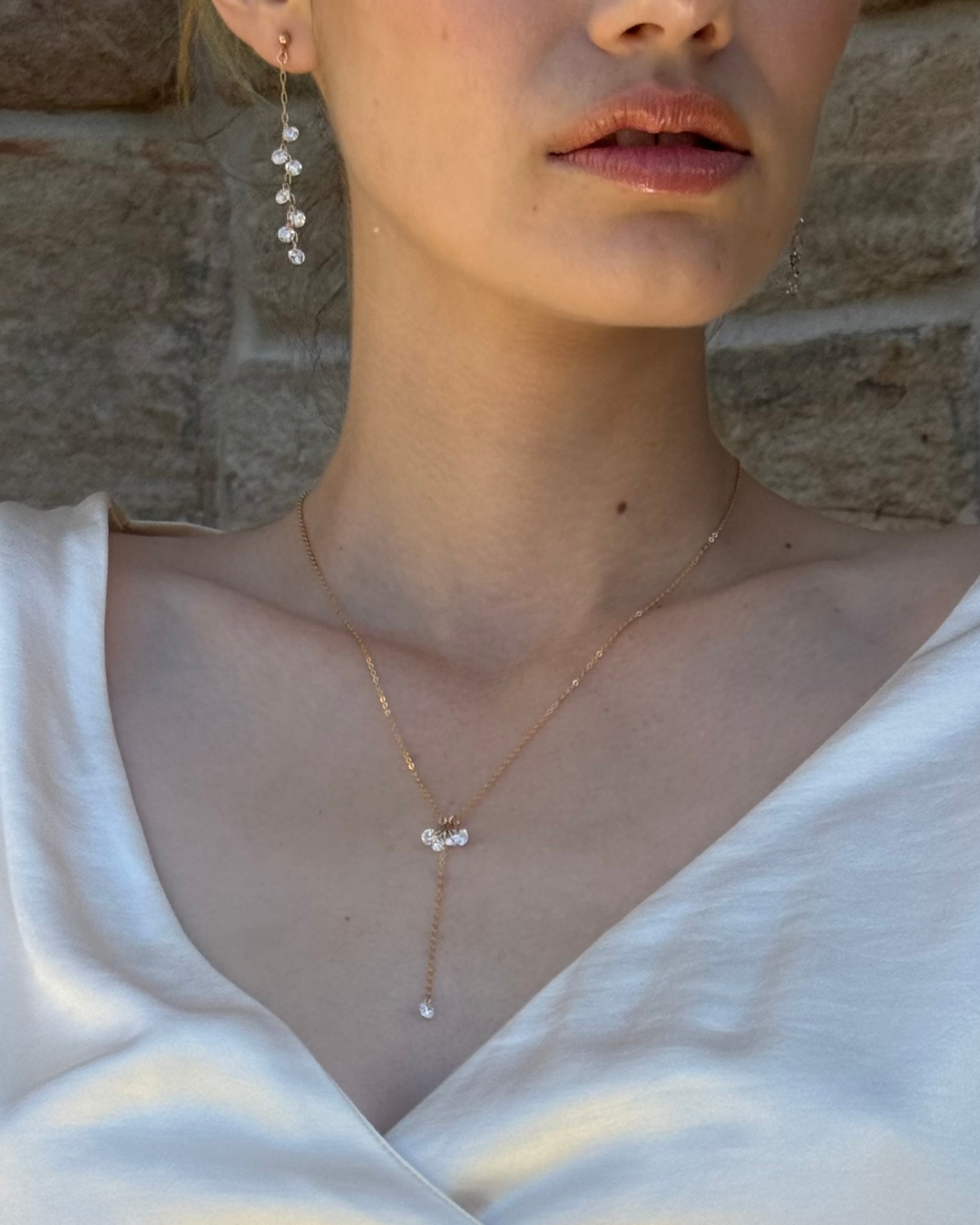 Close-up of a bride wearing a delicate necklace and earrings with cubic-zirconia against a stone wall background