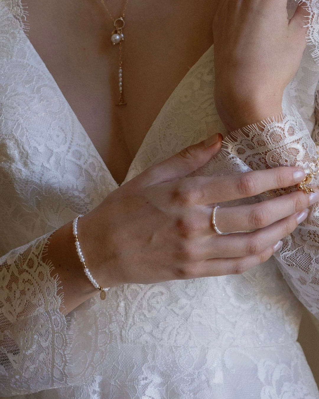 Close-up of a bride wearing gold and pearl jewellery including a necklace, ring, and bracelet on a white lace garment.