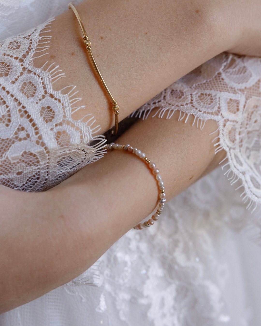 Close-up of a bride's arm wearing two gold and pearl bracelets with a lace garment in the background