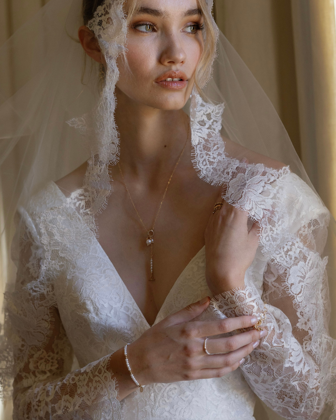 Woman wearing a lace wedding dress with a veil wearing gold and pearl jewellery.