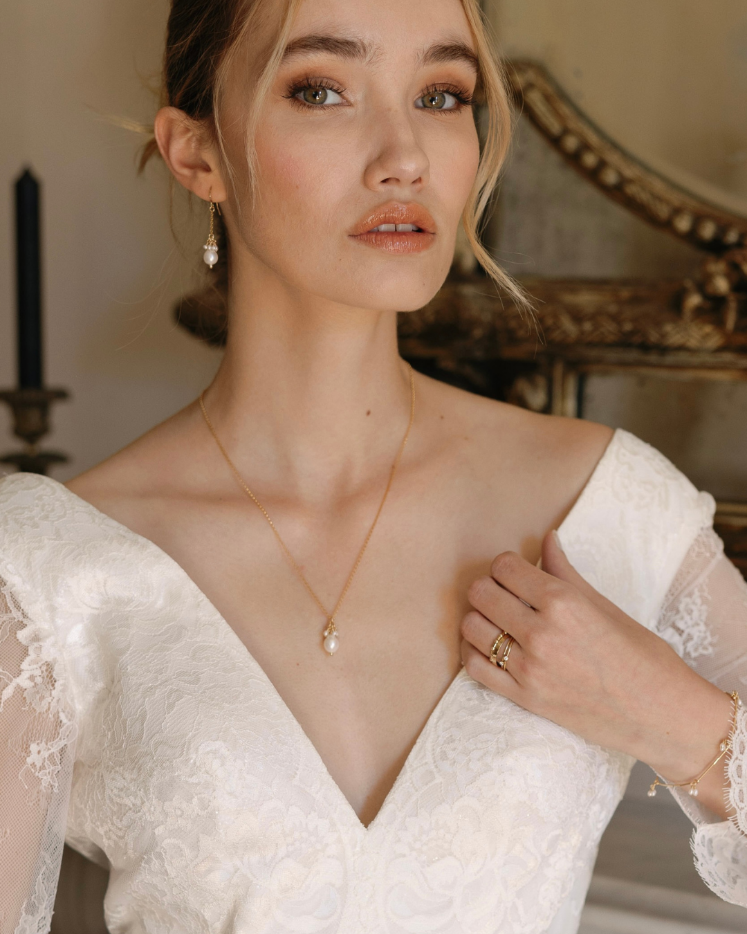 Woman wearing a white lace wedding dress with a deep neckline, gold and pearl necklace, and earrings.